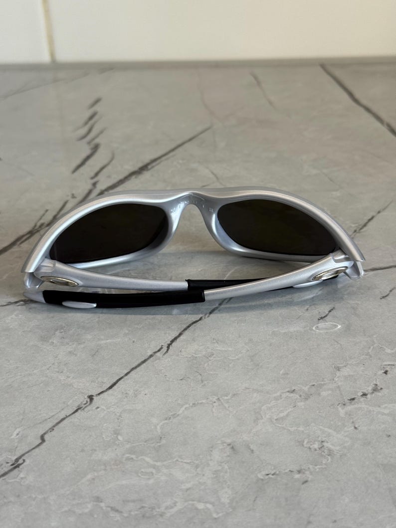 May include: Silver sunglasses with dark lenses. The sunglasses have a sleek, modern design with a curved frame and a black band at the temples. The lenses are dark, providing shade and protection from the sun.