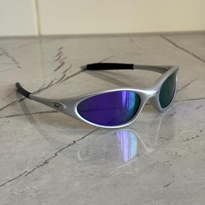 May include: Silver sunglasses with purple and green lenses. The sleek design features a curved frame and dark temples. The lenses reflect light, creating a vibrant effect. These sunglasses are a stylish accessory for sunny days.