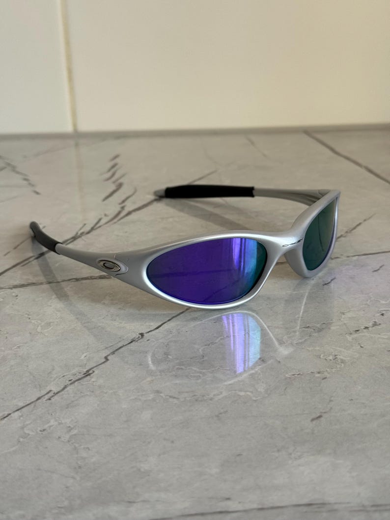 May include: Silver sunglasses with purple and green lenses. The sleek design features a curved frame and dark temples. The lenses reflect light, creating a vibrant effect. These sunglasses are a stylish accessory for sunny days.