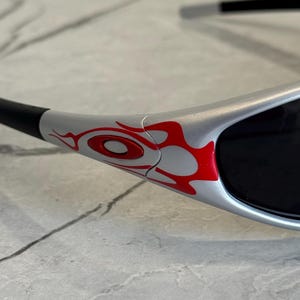 May include: Close-up of silver sunglasses with black lenses and arms. The frame features a red flame design on the side. The sunglasses are designed for eye protection and fashion.