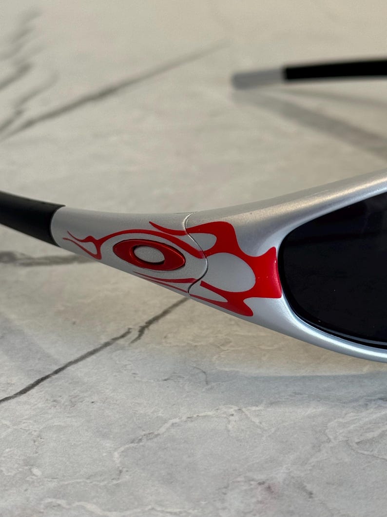 May include: Close-up of silver sunglasses with black lenses and arms. The frame features a red flame design on the side. The sunglasses are designed for eye protection and fashion.