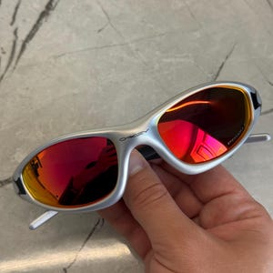 May include: Silver-framed sunglasses with red lenses. The sunglasses have a sleek, modern design with a curved frame and a prominent brand logo on the side. The lenses reflect a vibrant red hue.