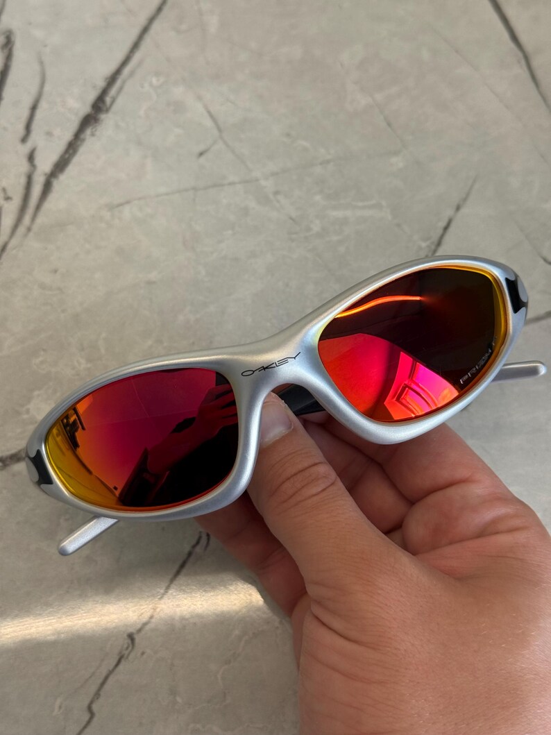 May include: Silver-framed sunglasses with red lenses. The sunglasses have a sleek, modern design with a curved frame and a prominent brand logo on the side. The lenses reflect a vibrant red hue.