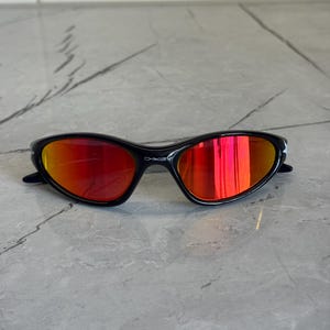 May include: Black sunglasses with red mirrored lenses. The sunglasses have a sleek, sporty design with a curved frame. The lenses reflect a vibrant orange and red hue. The brand name "Oakley" is visible on the frame.