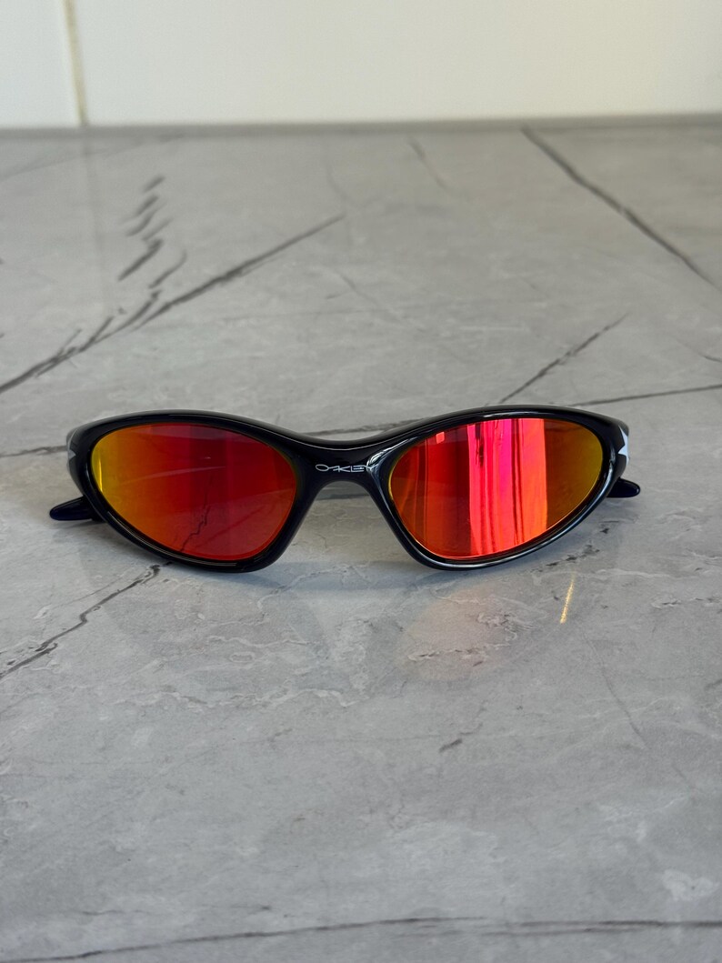 May include: Black sunglasses with red mirrored lenses. The sunglasses have a sleek, sporty design with a curved frame. The lenses reflect a vibrant orange and red hue. The brand name "Oakley" is visible on the frame.
