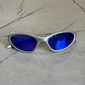May include: Silver sunglasses featuring striking blue lenses. The glasses have a streamlined, contemporary design with a curved frame and a small logo on the arm. The lenses reflect a vivid blue, providing a fashionable accessory.