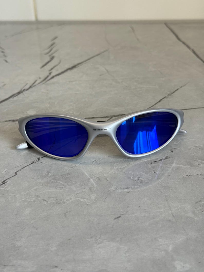 May include: Silver sunglasses featuring striking blue lenses. The glasses have a streamlined, contemporary design with a curved frame and a small logo on the arm. The lenses reflect a vivid blue, providing a fashionable accessory.