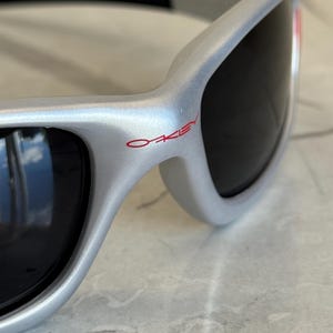 May include: Silver-framed sunglasses with black lenses. The brand name "OAKLEY" is printed in red on the frame. The sunglasses have a sleek, modern design, with a curved shape and a black arm.