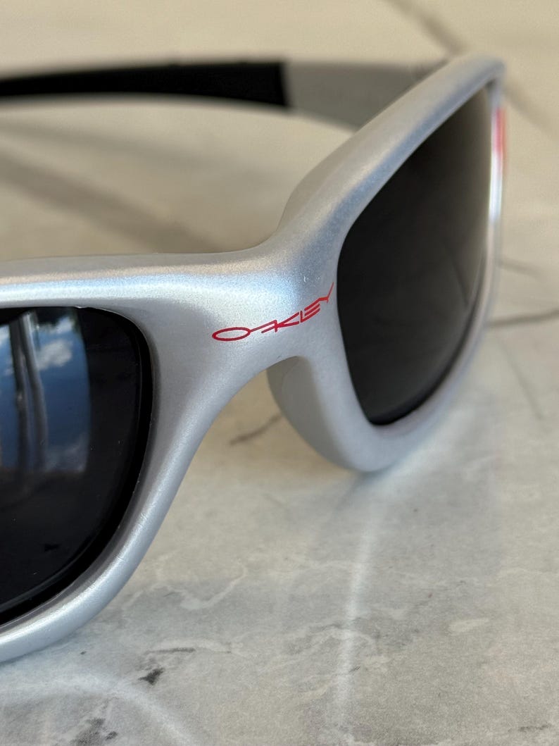 May include: Silver-framed sunglasses with black lenses. The brand name "OAKLEY" is printed in red on the frame. The sunglasses have a sleek, modern design, with a curved shape and a black arm.