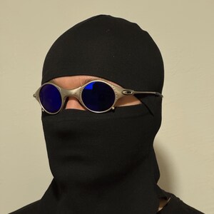 May include: Silver-framed sunglasses with blue lenses are worn over a black balaclava. The sunglasses have a rounded shape and a metallic finish. The balaclava covers the head and neck, leaving only the eyes visible.