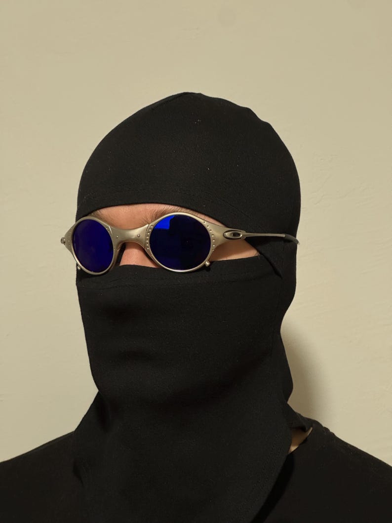May include: Silver-framed sunglasses with blue lenses are worn over a black balaclava. The sunglasses have a rounded shape and a metallic finish. The balaclava covers the head and neck, leaving only the eyes visible.