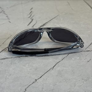 May include: Clear-framed sunglasses with dark lenses. The sunglasses have a sporty design with a curved bridge and temples. The lenses are a dark gray color, and the frame is transparent.