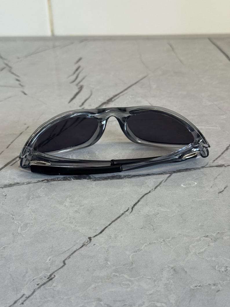 May include: Clear-framed sunglasses with dark lenses. The sunglasses have a sporty design with a curved bridge and temples. The lenses are a dark gray color, and the frame is transparent.