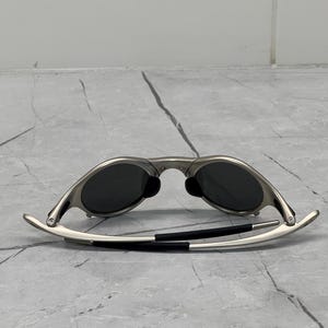 May include: Silver-framed sunglasses with dark lenses. The sunglasses have a sleek, modern design with curved temples and black accents. The lenses are oval-shaped and appear to be polarized.