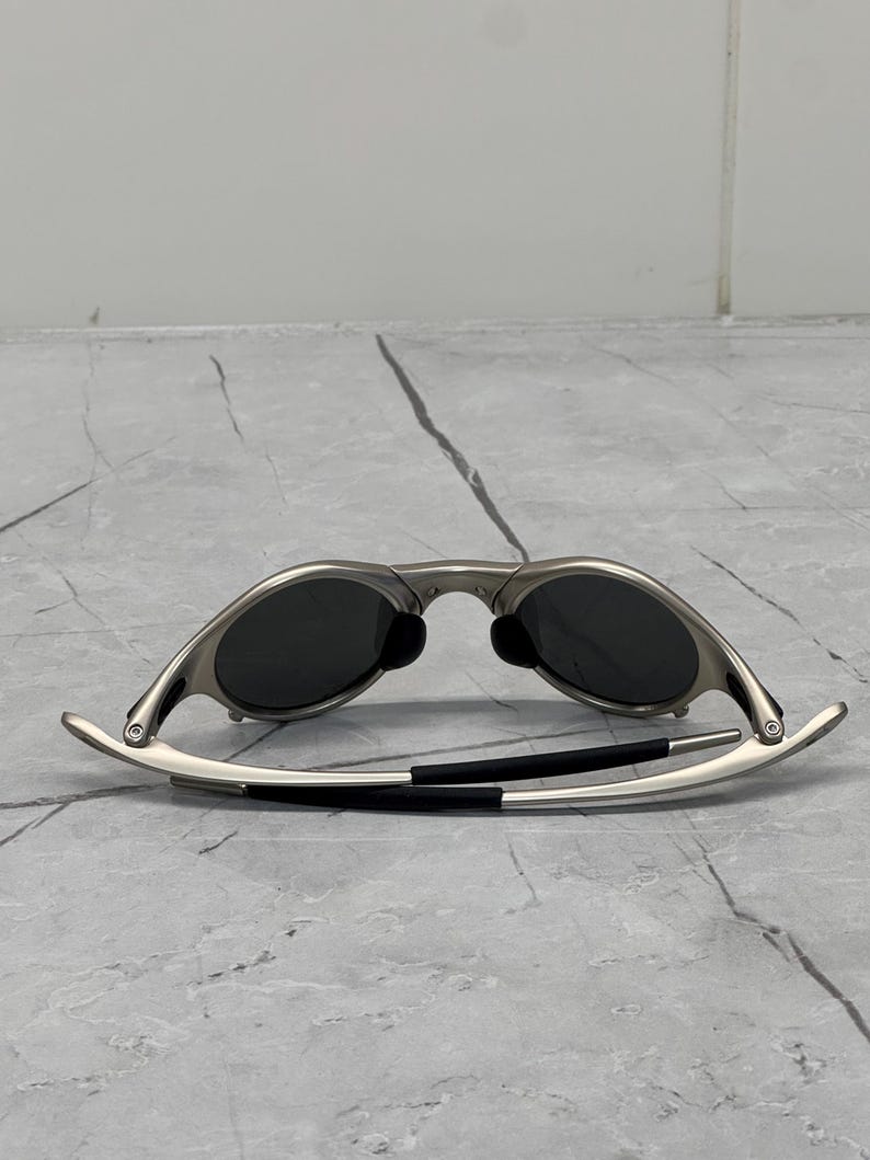 May include: Silver-framed sunglasses with dark lenses. The sunglasses have a sleek, modern design with curved temples and black accents. The lenses are oval-shaped and appear to be polarized.