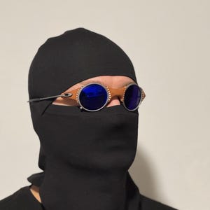 May include: A person wearing a black balaclava and a black shirt. The person is wearing round sunglasses with blue lenses and a brown frame.