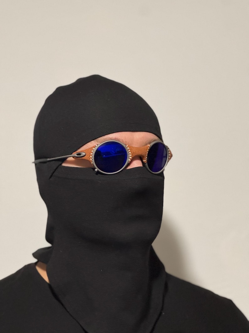 May include: A person wearing a black balaclava and a black shirt. The person is wearing round sunglasses with blue lenses and a brown frame.