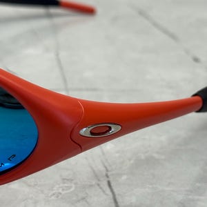 Op de afbeelding: Close-up van een sportieve zonnebril. De monturen zijn fel oranje, met een zilveren embleem aan de zijkant. De lenzen zijn reflecterend blauw. De veren zijn zwart en oranje.