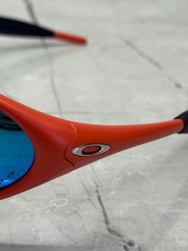 Op de afbeelding: Close-up van een sportieve zonnebril. De monturen zijn fel oranje, met een zilveren embleem aan de zijkant. De lenzen zijn reflecterend blauw. De veren zijn zwart en oranje.