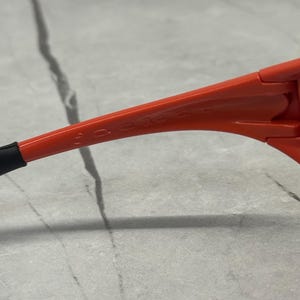 Op de afbeelding: Close-up van een oranje zonnebril. Het montuur is fel oranje, met de merknaam subtiel op de poot gedrukt. Het oortje is zwart. De zonnebril staat tegen een neutrale achtergrond.