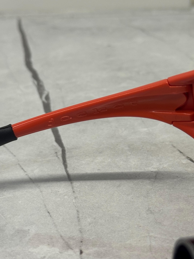 Op de afbeelding: Close-up van een oranje zonnebril. Het montuur is fel oranje, met de merknaam subtiel op de poot gedrukt. Het oortje is zwart. De zonnebril staat tegen een neutrale achtergrond.