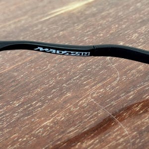 May include: Close-up of a black eyeglass frame arm with the word "MADMAN" printed in white. The frame is set against a wooden surface with a visible wood grain pattern.