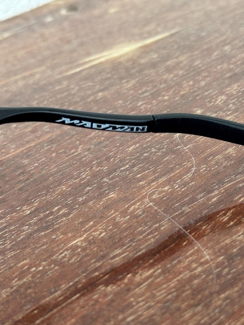 May include: Close-up of a black eyeglass frame arm with the word "MADMAN" printed in white. The frame is set against a wooden surface with a visible wood grain pattern.
