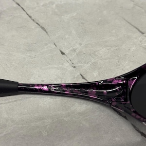 May include: Close-up of sunglasses with a black and purple marbled design. The sunglasses have a black arm and dark lenses. The brand name is printed in white on the arm of the sunglasses.
