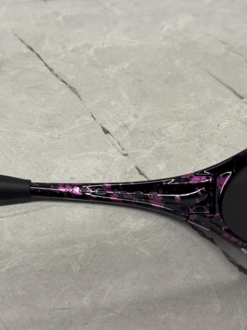 May include: Close-up of sunglasses with a black and purple marbled design. The sunglasses have a black arm and dark lenses. The brand name is printed in white on the arm of the sunglasses.