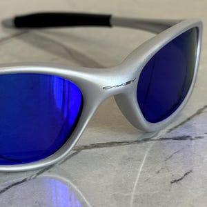 May include: Close-up of silver sunglasses with blue lenses. The sunglasses have a sleek, modern design with a black nose bridge and the brand name "Oakley" printed on the frame. The lenses reflect the surrounding environment.