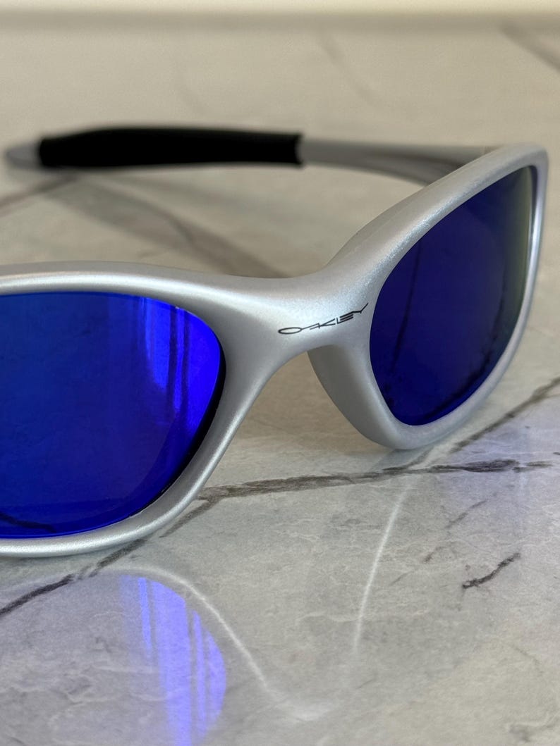 May include: Close-up of silver sunglasses with blue lenses. The sunglasses have a sleek, modern design with a black nose bridge and the brand name "Oakley" printed on the frame. The lenses reflect the surrounding environment.
