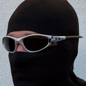 May include: Silver-framed sunglasses with dark lenses. The sunglasses have a decorative design on the temples. The person is wearing a black balaclava, covering the head and neck, with only the eyes visible.
