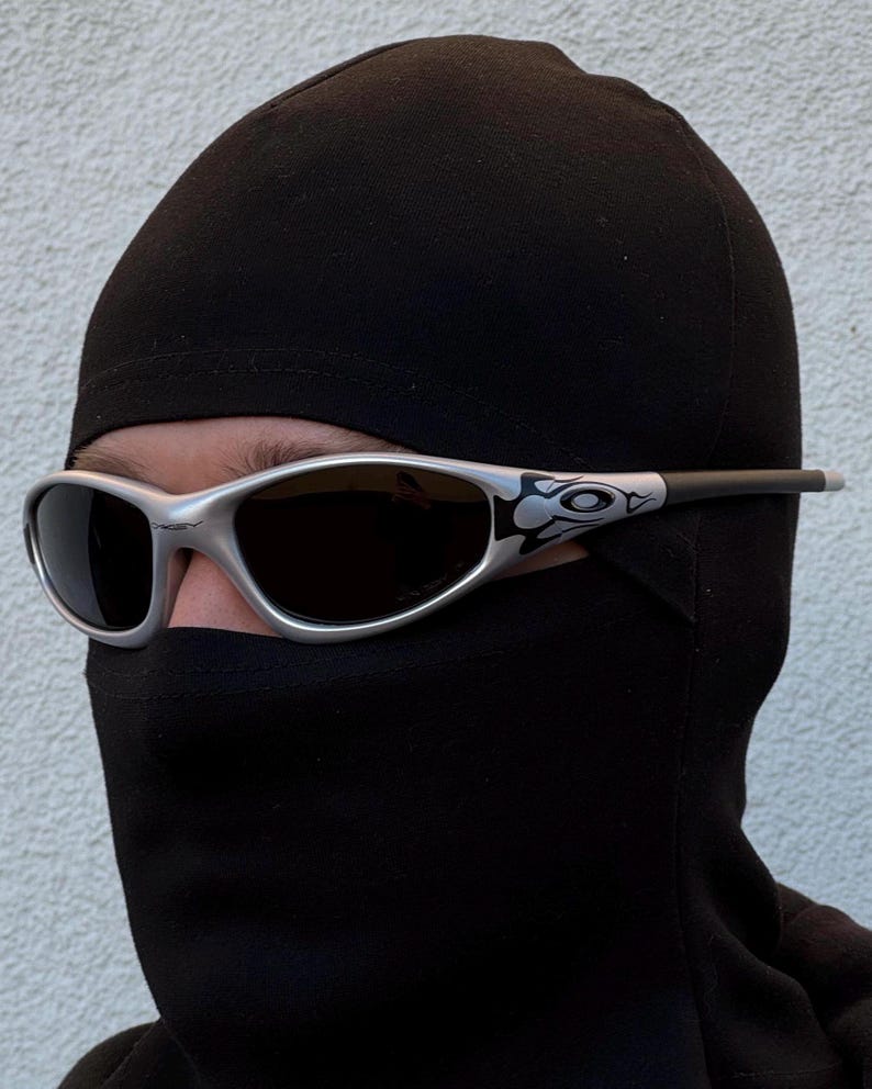 May include: Silver-framed sunglasses with dark lenses. The sunglasses have a decorative design on the temples. The person is wearing a black balaclava, covering the head and neck, with only the eyes visible.