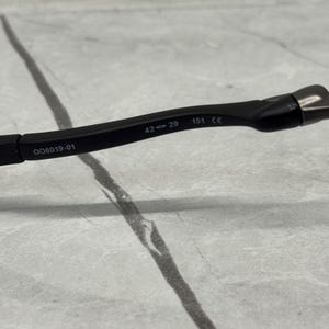 May include: Black eyeglasses frame with the numbers "42-29 151 CE" and the code "OO6019-01" printed on the arm. The frame is made of a matte black material.