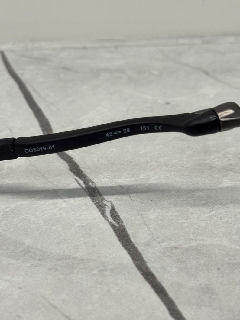May include: Black eyeglasses frame with the numbers "42-29 151 CE" and the code "OO6019-01" printed on the arm. The frame is made of a matte black material.