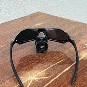Op de afbeelding: Zwarte zonnebril met een vergrootglas. De zonnebril heeft een strak ontwerp met donkere glazen en zwarte monturen. Het vergrootglas is gecentreerd tussen de glazen. De zonnebril ligt op een houten oppervlak.