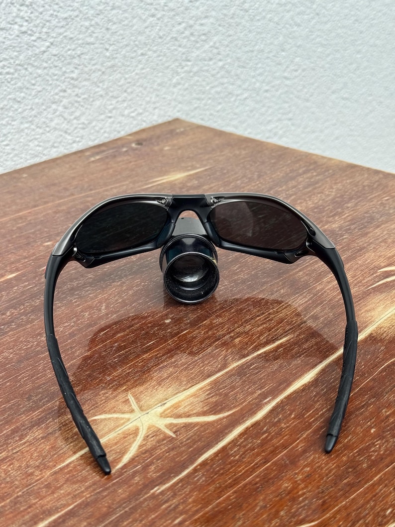 Op de afbeelding: Zwarte zonnebril met een vergrootglas. De zonnebril heeft een strak ontwerp met donkere glazen en zwarte monturen. Het vergrootglas is gecentreerd tussen de glazen. De zonnebril ligt op een houten oppervlak.