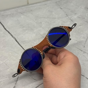 May include: Round sunglasses with blue lenses and a brown leather bridge. The frame is dark gray with a stitched leather detail on the sides. The lenses are a deep blue color, and the overall design is unique and stylish.