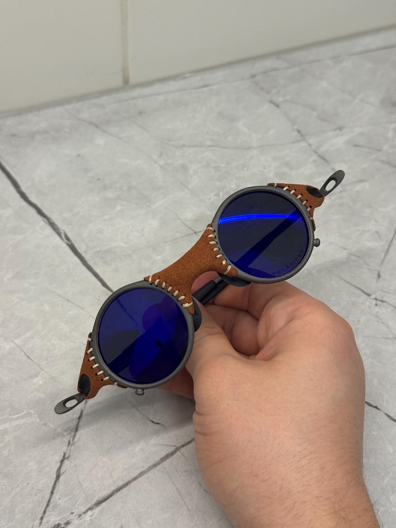 May include: Round sunglasses with blue lenses and a brown leather bridge. The frame is dark gray with a stitched leather detail on the sides. The lenses are a deep blue color, and the overall design is unique and stylish.