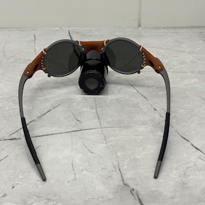 May include: Round sunglasses with dark lenses, a brown leather bridge, and gray metal frames. The leather is stitched to the frame. The sunglasses are resting on a black cylindrical object.
