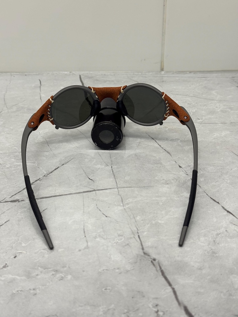 May include: Round sunglasses with dark lenses, a brown leather bridge, and gray metal frames. The leather is stitched to the frame. The sunglasses are resting on a black cylindrical object.