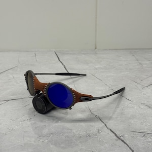 May include: Sunglasses with a brown leather and black frame. The lenses are a vibrant blue. The frame has a unique design with visible stitching. The sunglasses are resting on a gray surface.
