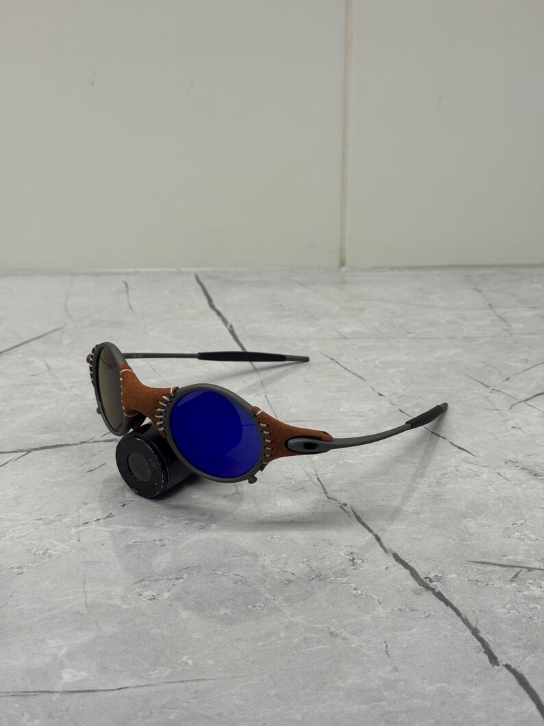 May include: Sunglasses with a brown leather and black frame. The lenses are a vibrant blue. The frame has a unique design with visible stitching. The sunglasses are resting on a gray surface.