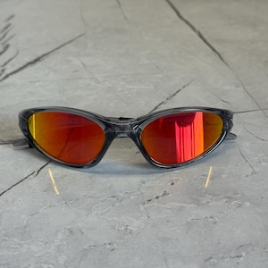 K&ouml;nnte beinhalten: Sportsonnenbrille mit grauem Rahmen und leuchtend roten verspiegelten Gl&auml;sern. Die Sonnenbrille hat ein schlankes, modernes Design, das sich f&uuml;r Outdoor-Aktivit&auml;ten eignet. Die Gl&auml;ser reflektieren einen hellen Orangeton.