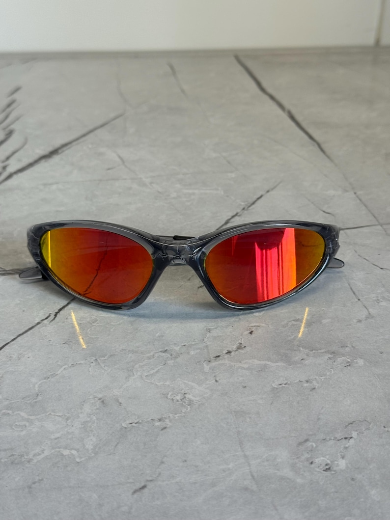 K&ouml;nnte beinhalten: Sportsonnenbrille mit grauem Rahmen und leuchtend roten verspiegelten Gl&auml;sern. Die Sonnenbrille hat ein schlankes, modernes Design, das sich f&uuml;r Outdoor-Aktivit&auml;ten eignet. Die Gl&auml;ser reflektieren einen hellen Orangeton.