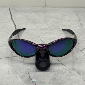 May include: Sunglasses with a black and purple speckled frame and mirrored lenses. The lenses reflect a blue and green hue. The text "BRAIN DEAD" is visible on the frame. The sunglasses are resting on a black cylindrical object.