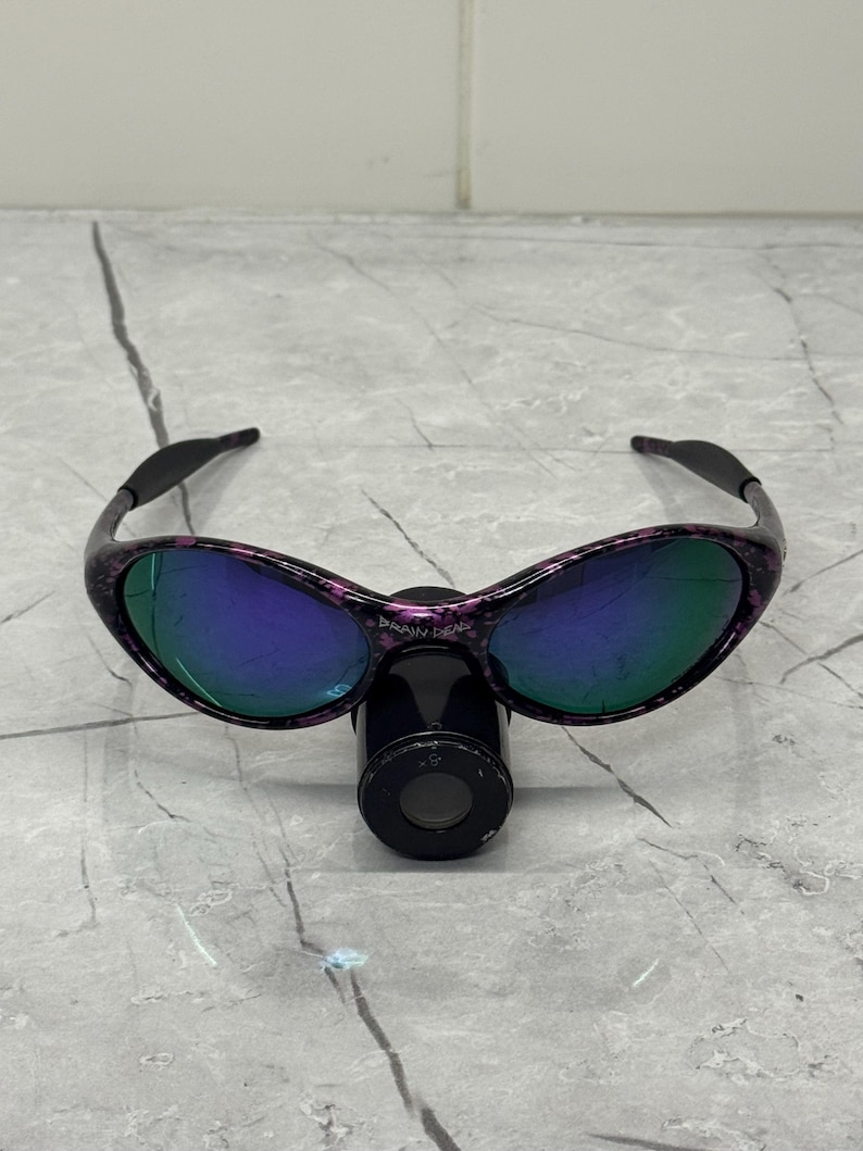 May include: Sunglasses with a black and purple speckled frame and mirrored lenses. The lenses reflect a blue and green hue. The text "BRAIN DEAD" is visible on the frame. The sunglasses are resting on a black cylindrical object.