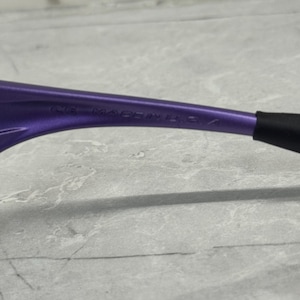 May include: Close-up of a purple eyeglass frame with the text "CE MADE IN U.S.A." The frame has a sleek, modern design with a black arm.