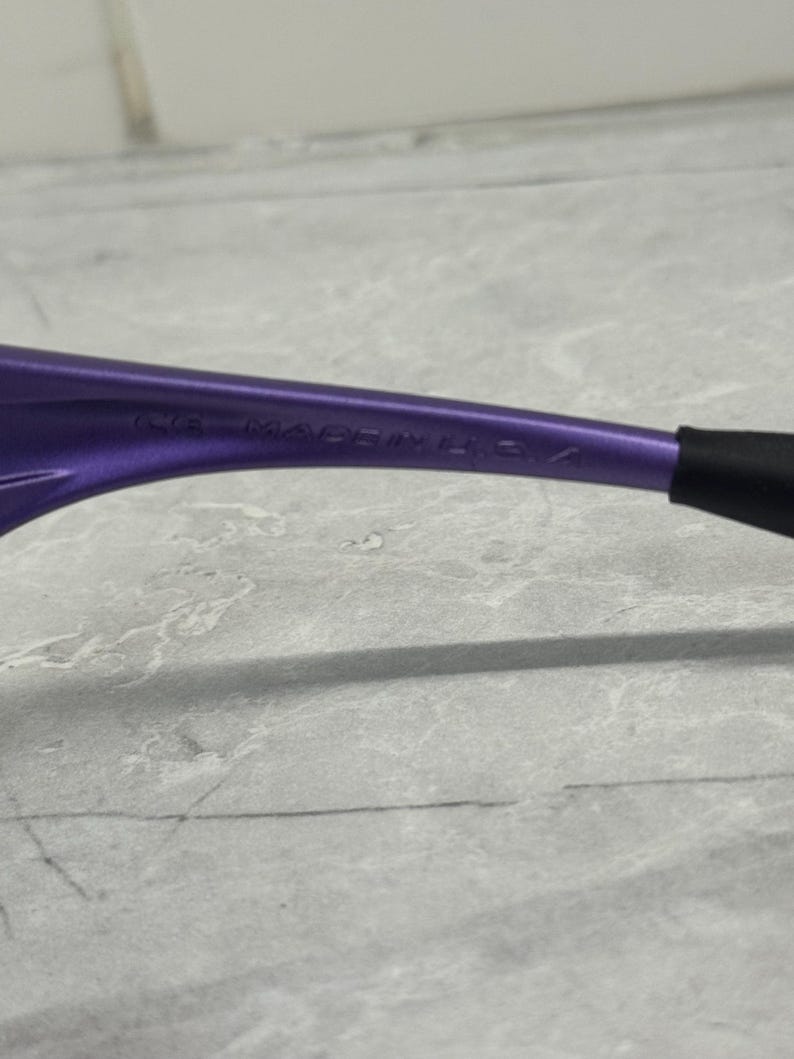 May include: Close-up of a purple eyeglass frame with the text "CE MADE IN U.S.A." The frame has a sleek, modern design with a black arm.