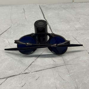 May include: Black sunglasses with blue lenses are displayed on a black cylindrical object. The sunglasses have a sleek, modern design with thin temples and a bridge. The lenses are a vibrant blue color.